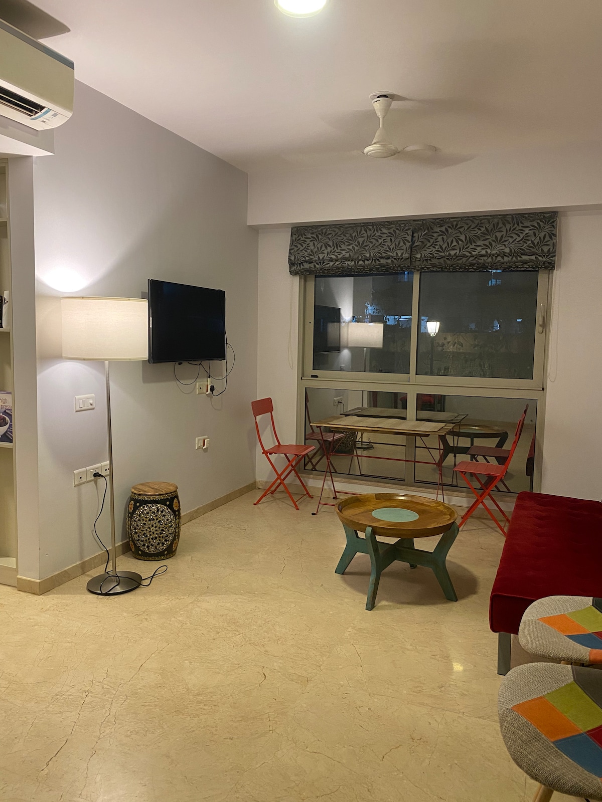 The living area features a sleek television mounted on the wall, with a comfortable seating arrangement that includes a red seating unit and colorful stools. A small table sits in the center, surrounded by two bright red chairs, with soft lighting provided by a floor lamp.
