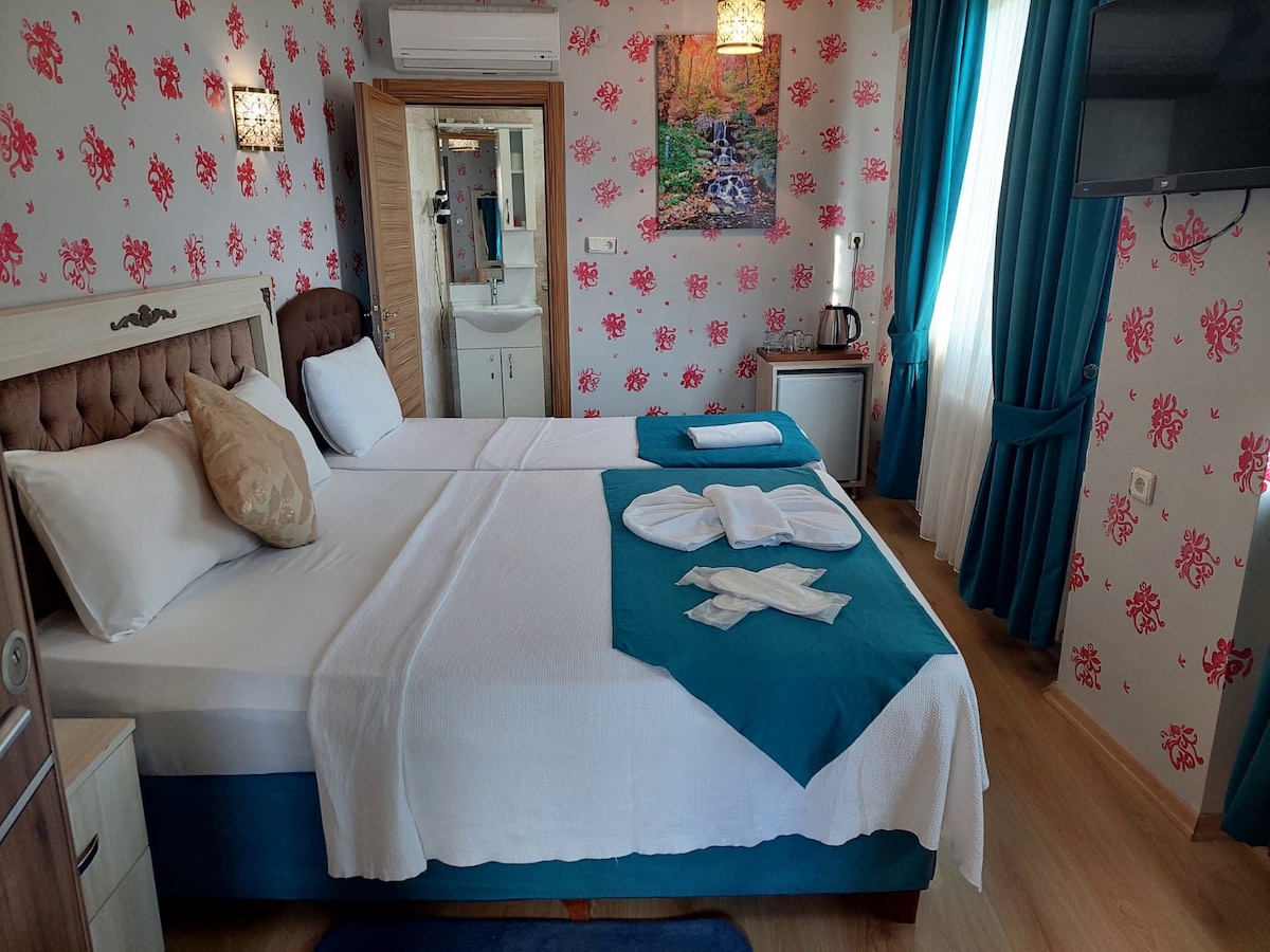 A cozy guest room features a spacious bed with a light blue accent blanket, complemented by neatly arranged towels. Floral wallpaper adds a soft touch, while a small kitchenette and a flat-screen TV are positioned nearby. Natural light filters through the blue curtains.