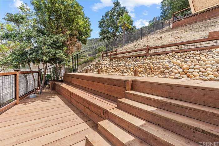 A wooden deck features multiple steps leading up a landscaped area, where smooth stones are arranged along the slope. Lush greenery adds a natural element to the setting, and a clear blue sky is visible above.