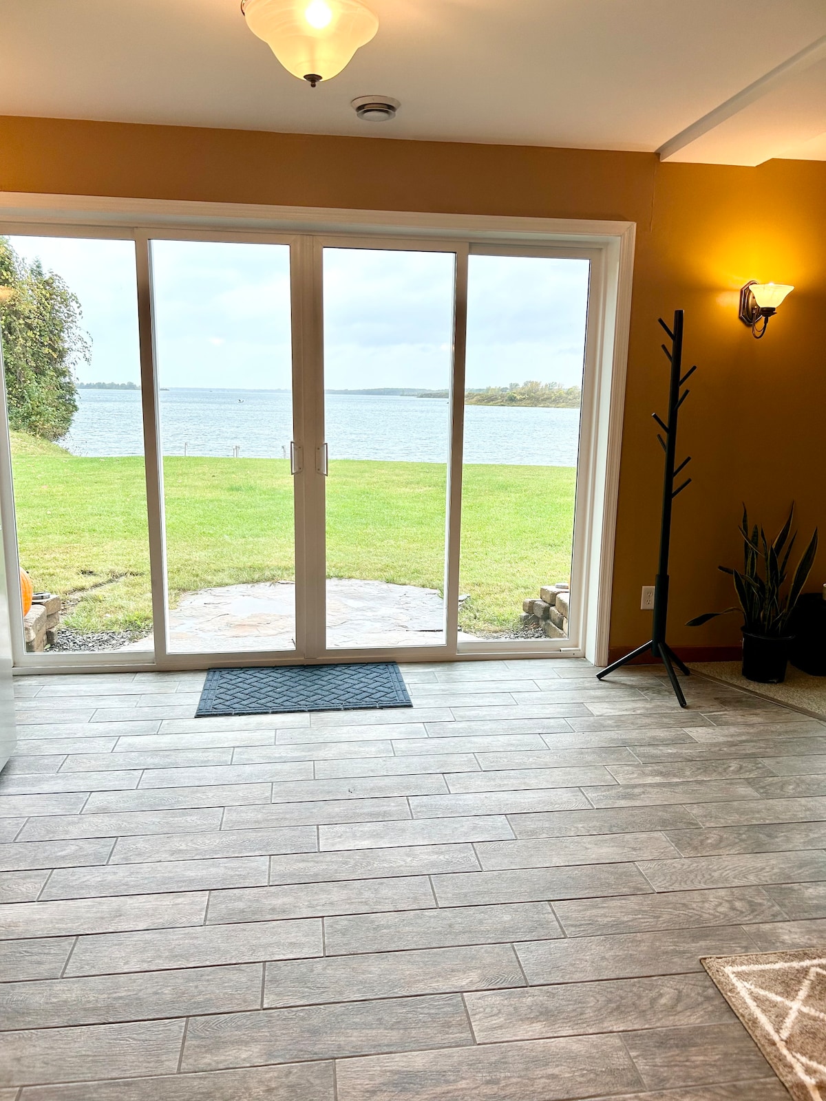 A well-lit entryway features sliding glass doors that open to a grassy area overlooking the water. Natural light fills the space, highlighting the tile flooring. A coat rack stands to one side, while potted plants add a touch of greenery.