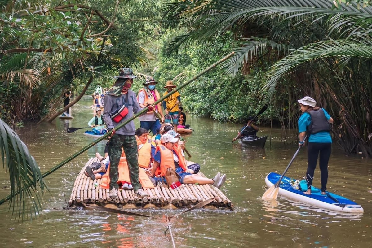 A tranquil scene of kayakers and a bamboo raft navigating through a lush mangrove waterway. Dense green foliage frames the water while individuals in life jackets engage in the serene activity. The environment showcases a blend of nature and local exploration.