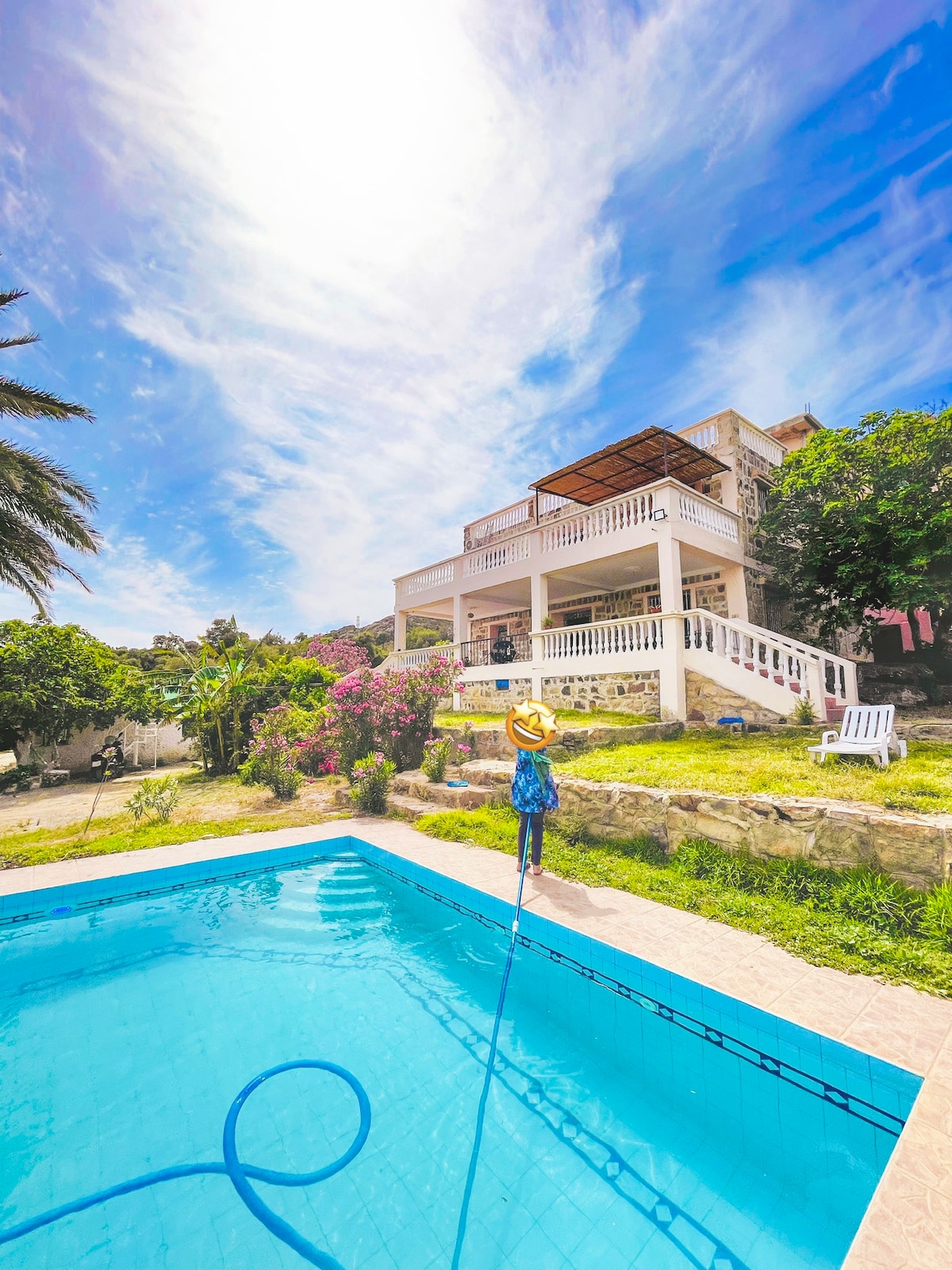 A refreshing swimming pool is framed by well-maintained landscaping, featuring vibrant flowers and palm trees. The multilevel retreat is visible in the background, with a spacious terrace and white railings under a bright sky. Sunlight reflects off the pool's clear water.