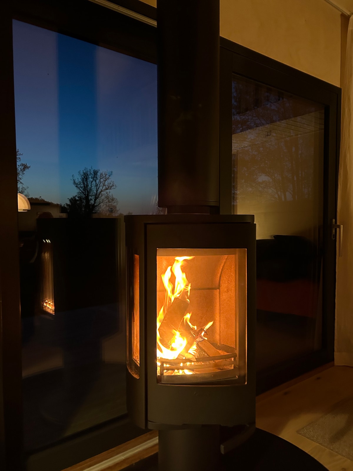 A modern wood stove emits a warm glow, with flames visibly dancing inside its glass enclosure. The background features large glass doors, revealing an evening sky transitioning from light blue to darker hues, creating a cozy indoor atmosphere.