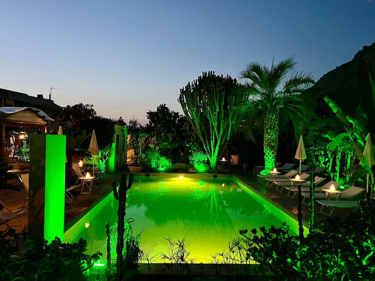 An outdoor pool is surrounded by lush greenery, with vibrant lights casting a green hue. Sun loungers are arranged nearby under umbrellas, enhancing the serene atmosphere. The scene is set against a twilight sky, adding to the tranquil setting of the garden.