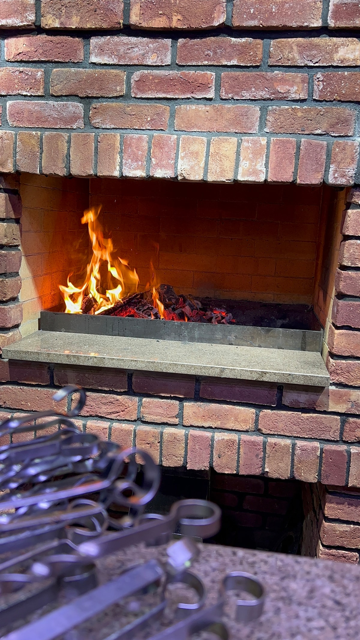 A traditional brick fireplace is seen, featuring flames dancing above glowing embers. Cooking utensils are placed in the foreground, adding context to the setting. The warm tones of the bricks contrast with the bright fire, creating an inviting focal point.