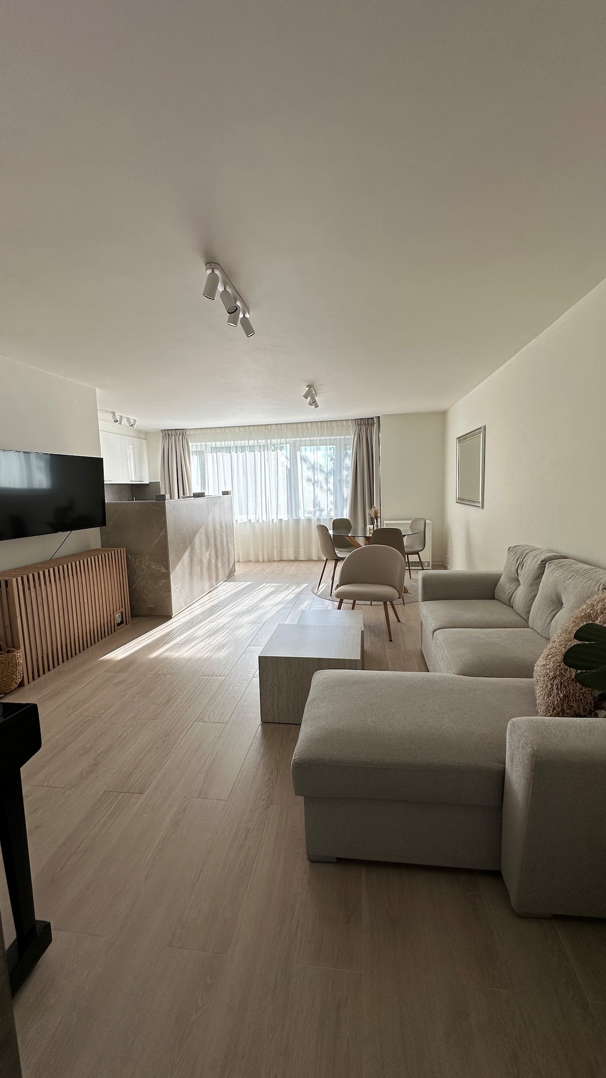 A modern living area features a light color palette, with a comfortable sofa arrangement and a coffee table. Natural light floods the space through large windows, complemented by minimalist decor and a television positioned against the wall, creating a spacious and inviting atmosphere.