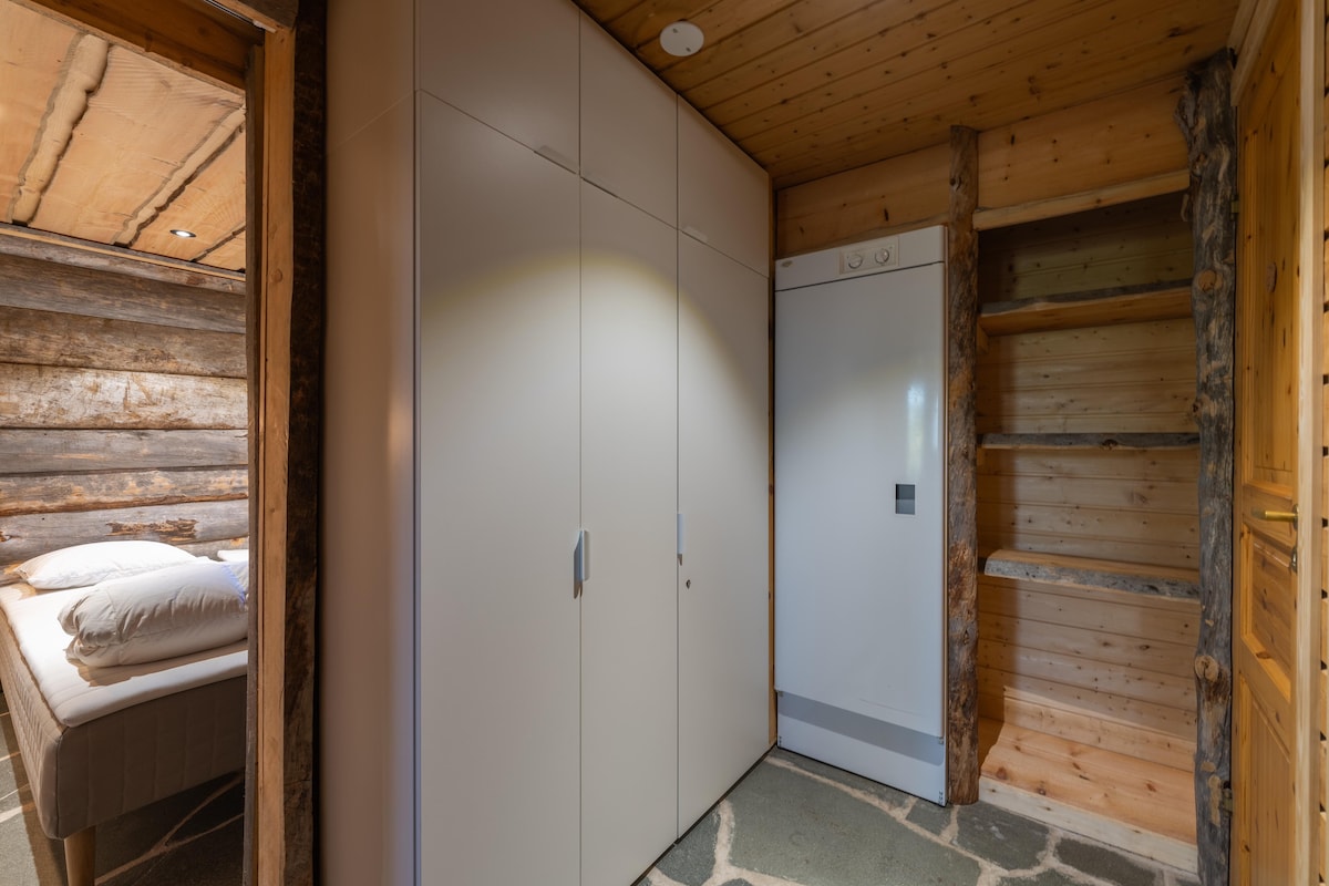 A practical entryway features modern, sleek storage cabinets alongside a refrigerator. The rustic log structure of the house adds warmth to the space. Soft lighting highlights the clean lines of the cabinets, while access to a cozy sleeping area is visible in the background.