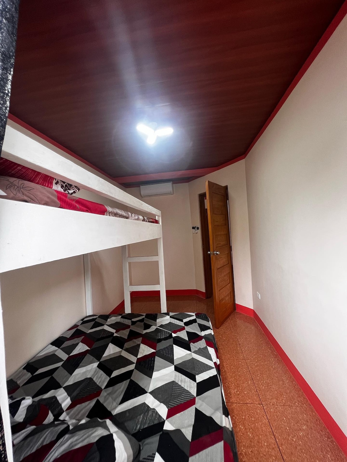 A cozy sleeping area is presented, featuring a double-deck bed with a patterned comforter. The well-lit space showcases a warm wooden ceiling and a simple door. Additional floor space is available, allowing for easy movement.