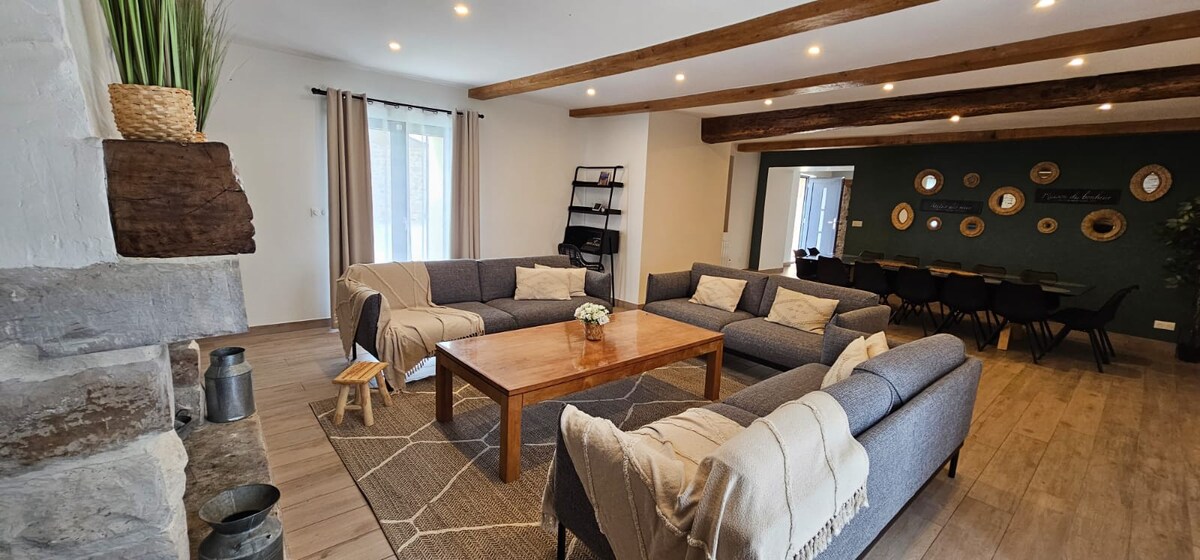 A spacious living area features multiple grey sofas arranged around a central wooden coffee table. Soft lighting illuminates the room, while wooden beams overhead add character. Decorative wall mirrors and a small bookshelf complete the inviting atmosphere.