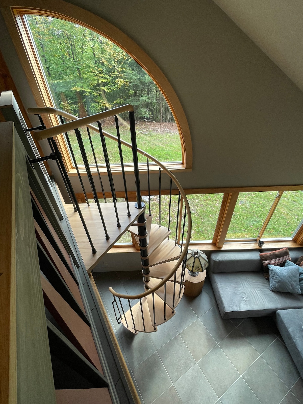 A circular staircase extends from the loft area to the lower level, showcasing a blend of wood and metal. Large windows frame the scenic view of surrounding trees, while a comfortable seating area is positioned nearby, enhancing the studio's welcoming ambiance.