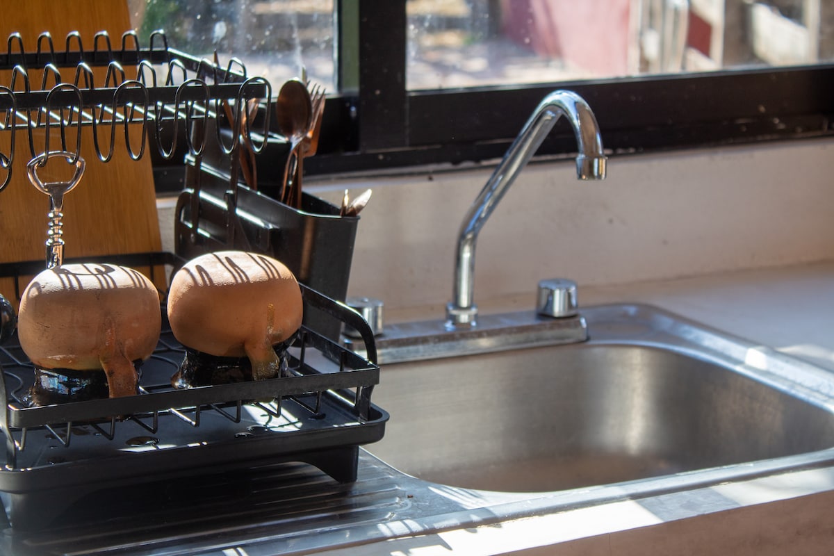 A kitchen counter is shown with a sleek stainless steel sink. A black dish rack holds two decorated clay pots alongside utensils, creating a functional and organized space. Natural light streams in through a nearby window, enhancing the cheerful ambiance.