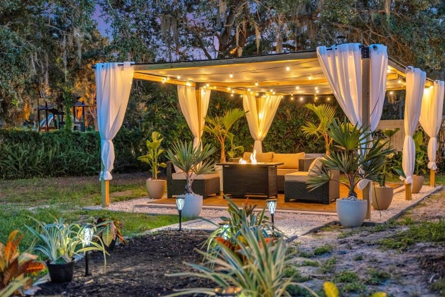 An outdoor pergola is illuminated with string lights, creating a warm ambiance. Comfortable seating is provided with decorative cushions surrounded by potted plants. Soft landscaping features colorful plants and pathway lighting, enhancing the tranquil outdoor space.