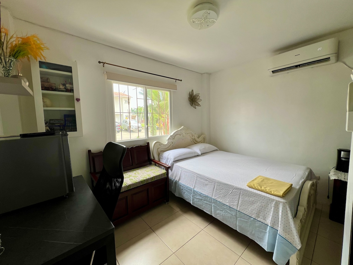 A well-lit bedroom features a spacious bed with light bedding, complemented by a decorative wall accent. A small desk and chair provide a workspace, while a cozy seating area by the window adds comfort. Natural light fills the room through a large window, enhancing the serene atmosphere.