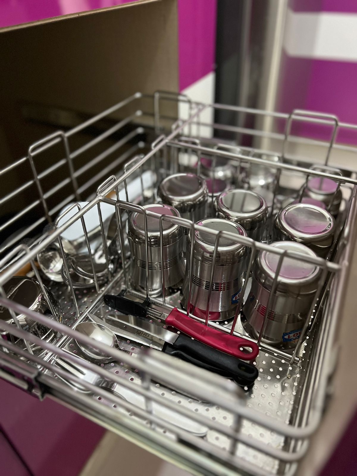 A stainless steel dish rack is filled with a variety of kitchen items, including several metal cups, cutlery, and utensils. The vibrant purple wall in the background adds a pop of color to the modern setting.