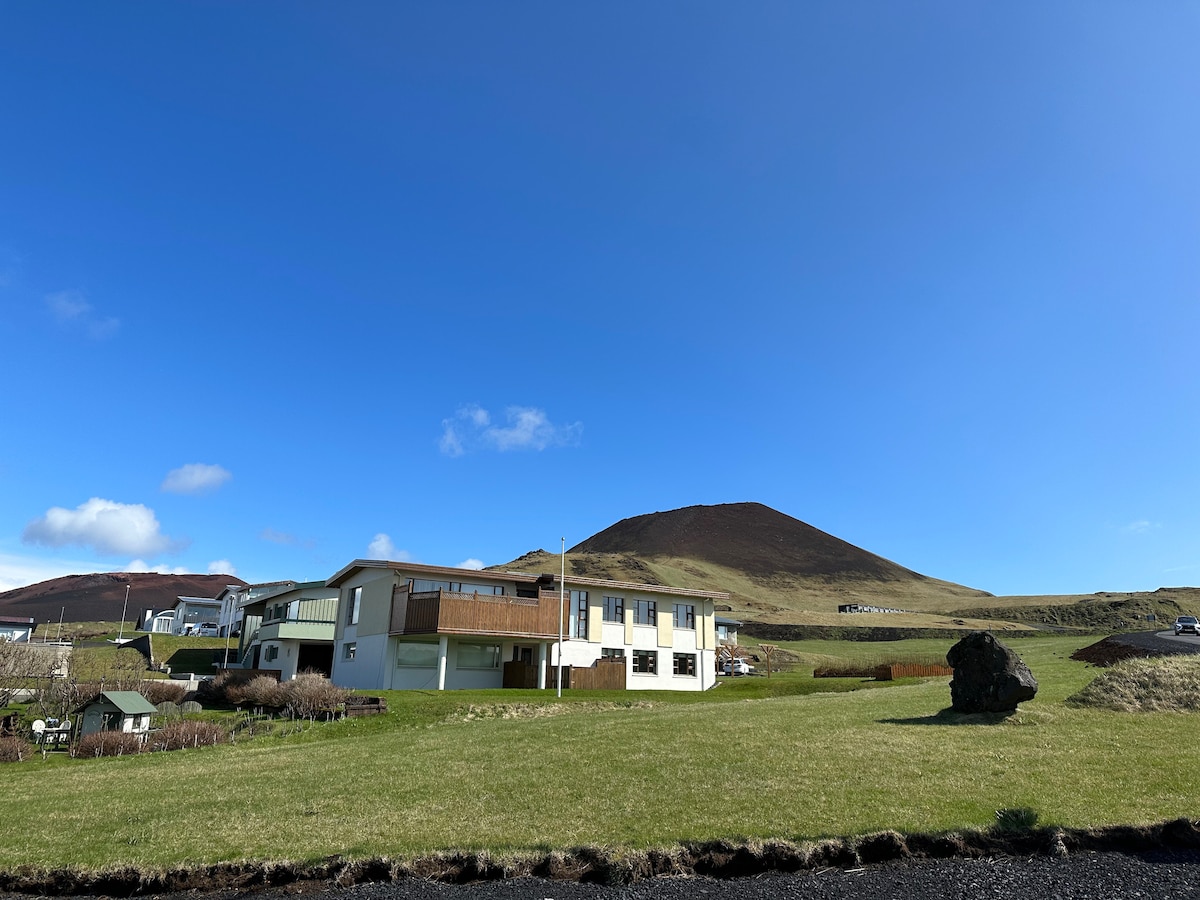 A well-structured building is set against a backdrop of rolling green hills and a volcanic mountain. The clear blue sky enhances the serene environment, while the landscaped grass area features scattered rocks, adding to the natural charm of the setting.
