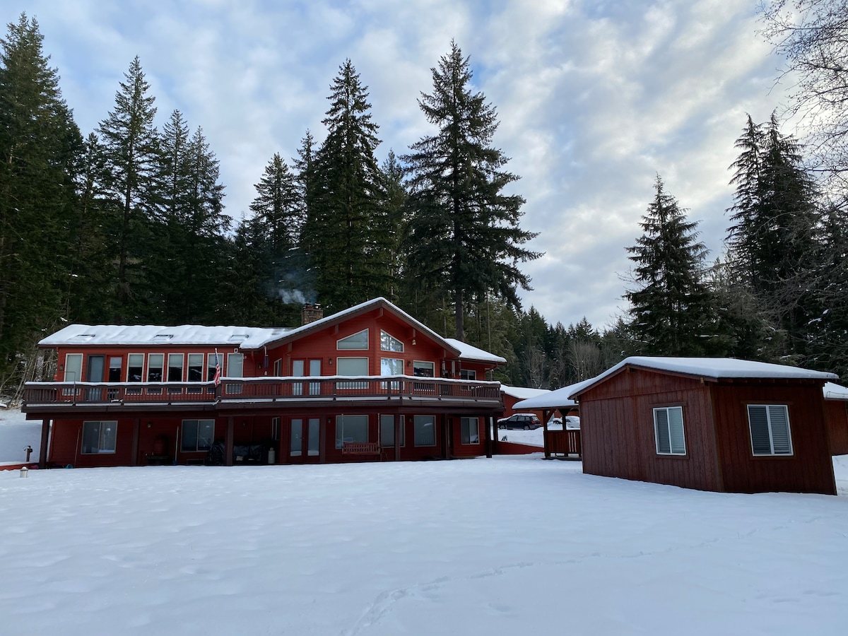 A two-story red cabin is surrounded by a snowy landscape, with tall evergreen trees in the background. The structure features multiple balconies and large windows, creating a spacious outdoor area covered in fresh snow.