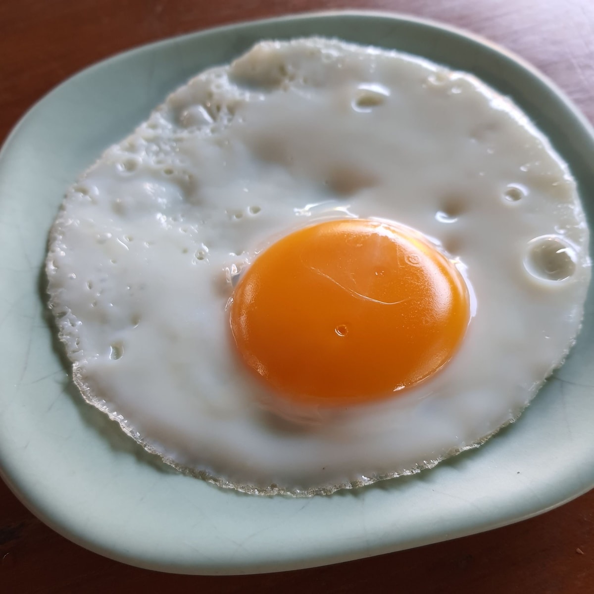 A single fried egg is displayed on a light green ceramic plate. The egg white has a slightly crisp edge, while the vibrant orange yolk is centered and glossy. The natural wood surface beneath the plate provides a warm contrast.