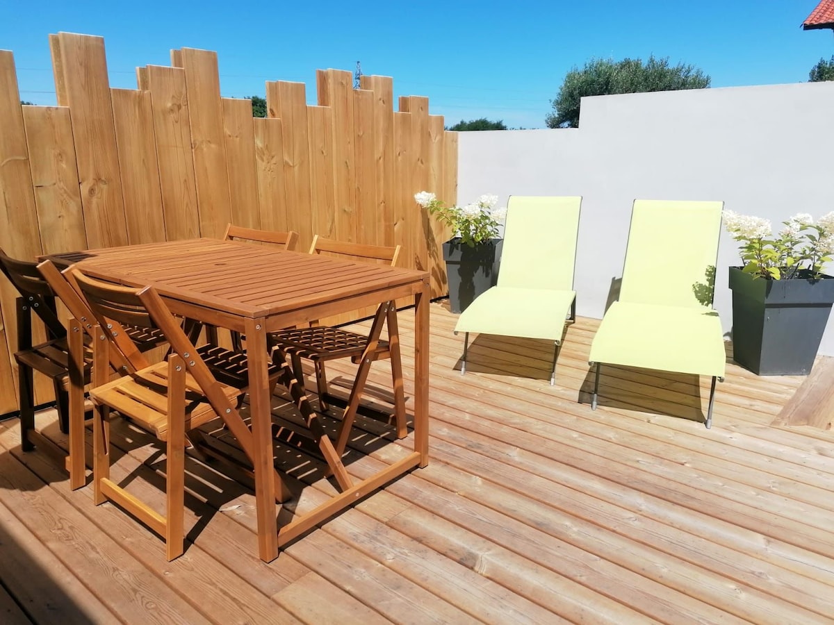 An independent terrace is featured, furnished with a wooden dining table and six chairs. Two lounge chairs in a light green color are positioned beside potted plants, all set against a backdrop of wooden fencing and clear blue skies.