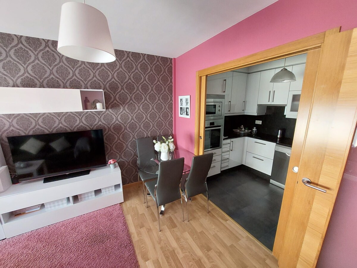 A cozy living area is shown with a large television against a patterned pink wall. A small dining table with four chairs is set near a doorway leading to a modern kitchen with sleek appliances and cabinetry, providing a sense of openness.