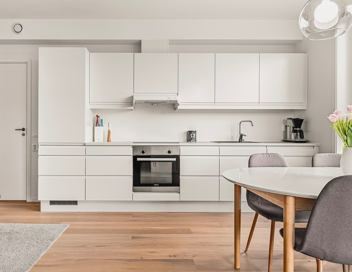 A modern kitchen space features sleek white cabinetry and a stainless steel oven. A circular dining table with gray chairs is positioned nearby, while a small rug adds texture to the wooden floor. Soft lighting enhances the clean, minimalist design.