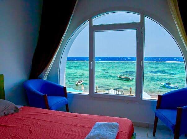 A large circular window frames a stunning view of the sea, with clear turquoise waters visible just outside. Two blue chairs sit beside the window. The sleeping area features a spacious bed with a simple bedspread, creating a serene setting enhanced by natural light.