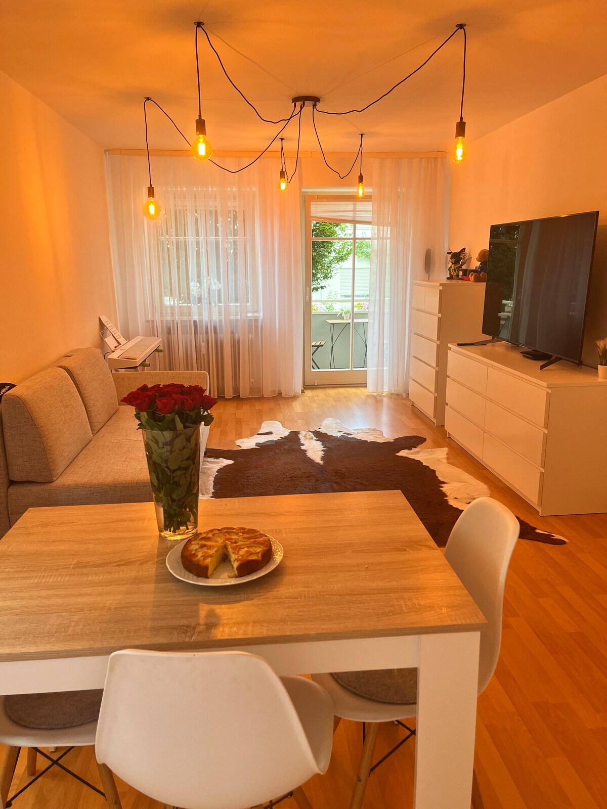 A spacious living area features a comfortable sofa, accompanied by a dining table with modern chairs. A large television is mounted on the wall, while natural light filters through sheer curtains. A decorative table holds a plate of pastries, enhancing the warm ambiance.