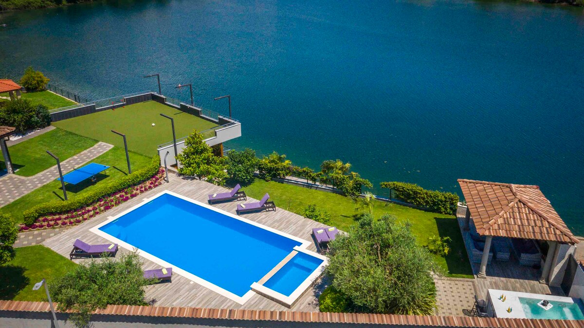 An aerial view captures a serene outdoor area featuring a large blue swimming pool surrounded by wooden decking. Sun loungers in purple are arranged thoughtfully around the pool, complemented by grassy spaces and tropical greenery alongside the tranquil water.