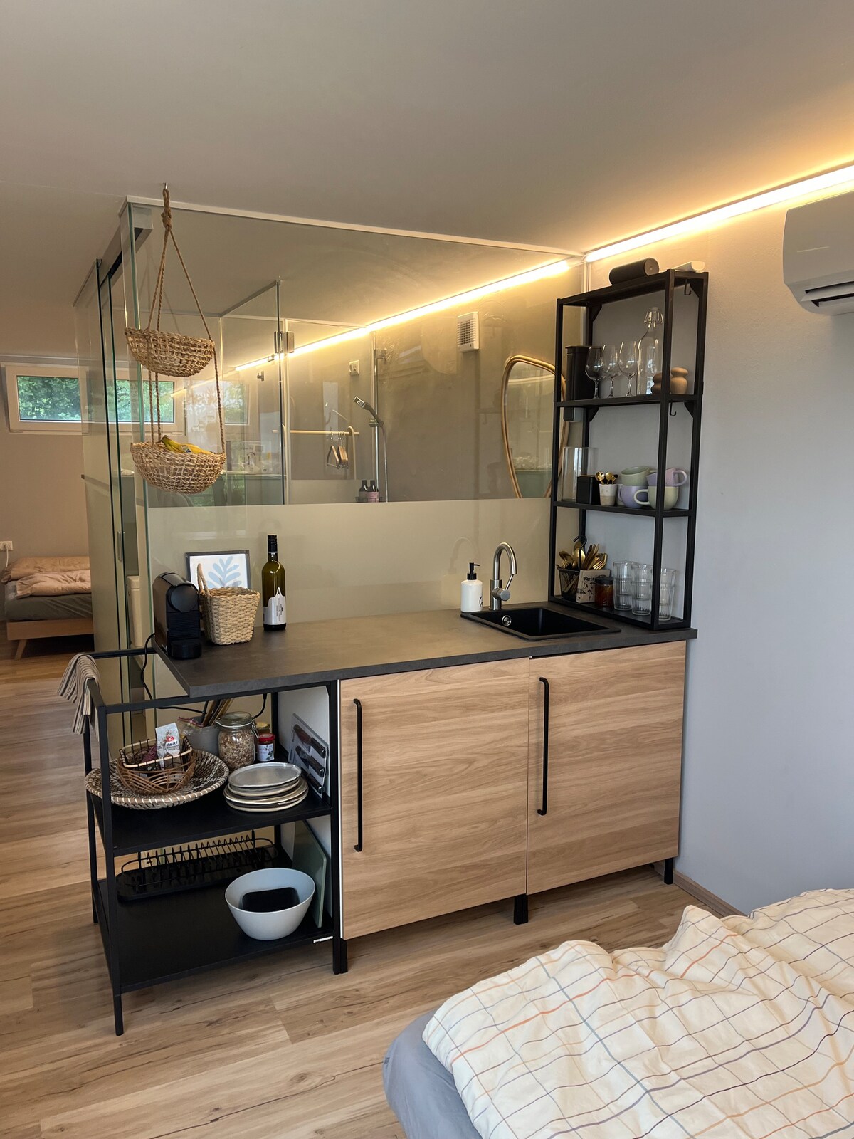 A modern kitchenette is displayed, featuring wooden cabinets with black handles and a sleek countertop. Open shelving holds glassware and decorative items. A small sink is integrated, while a cozy sleeping area is visible in the background, enhancing the space's functionality.