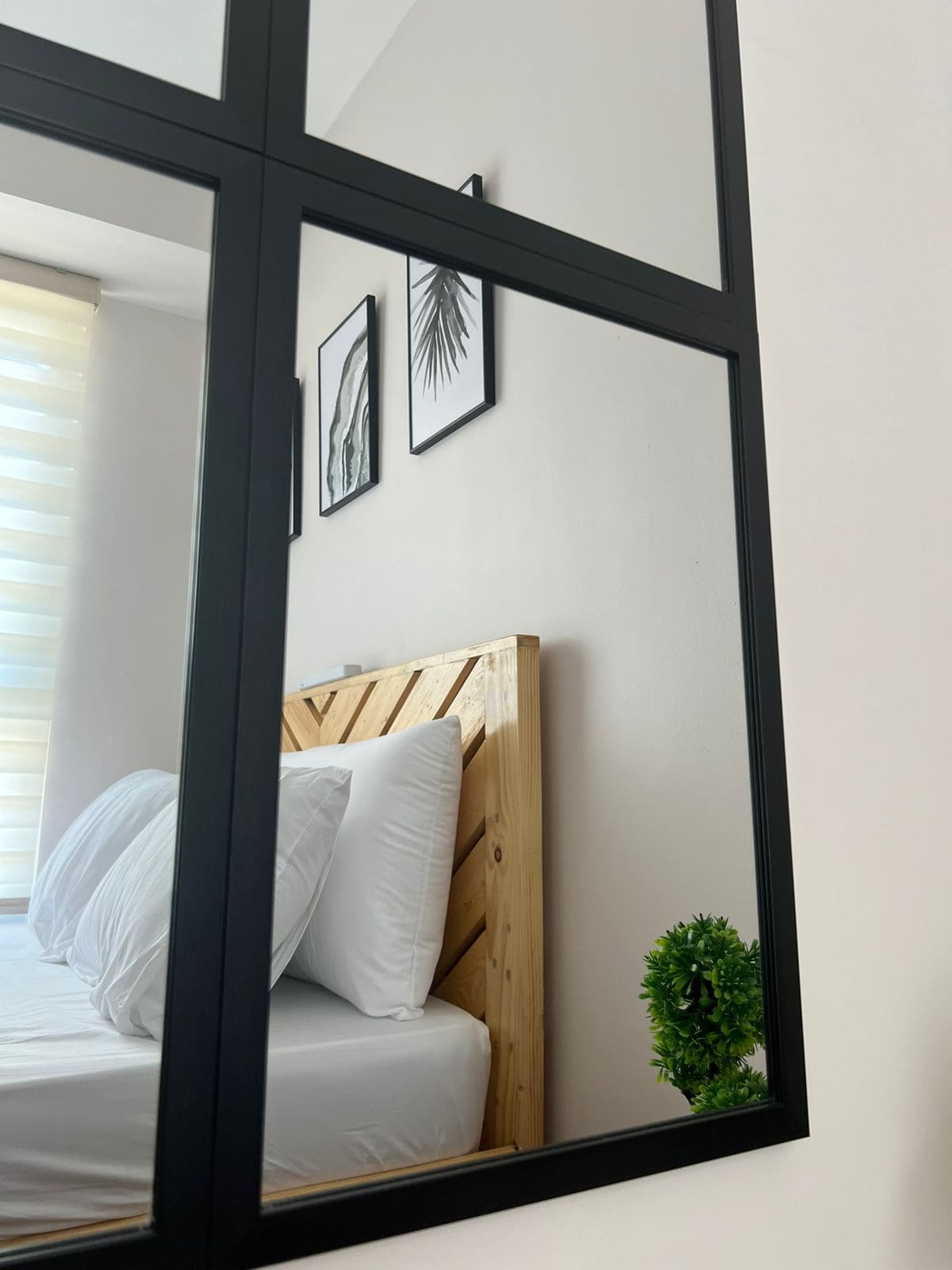 A mirror reflects a cozy sleeping area featuring a wooden headboard with decorative wall art above it. Light-colored bedding enhances the serene ambiance, while a small green plant provides a touch of nature to the space. Window treatments allow soft light to filter in.