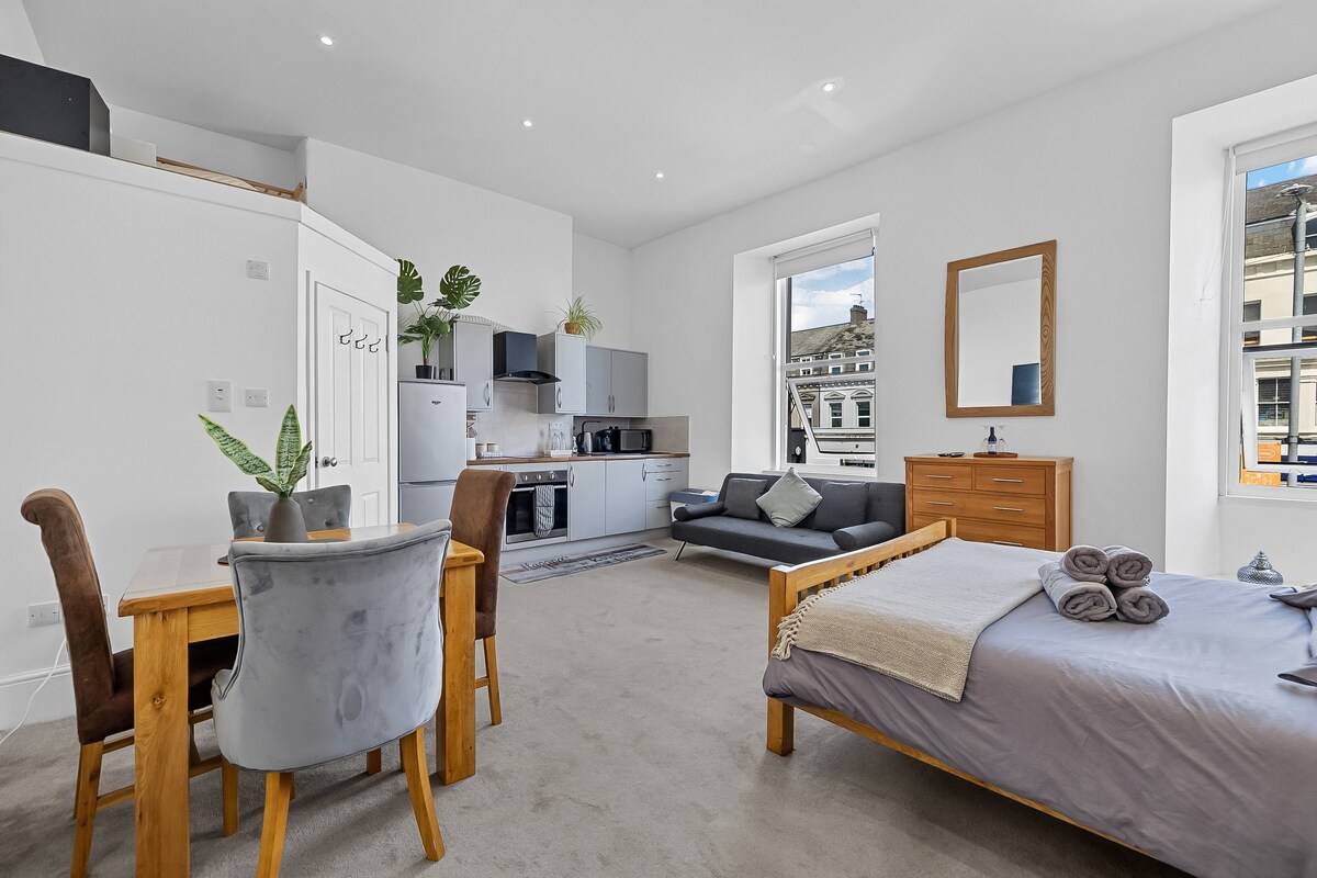 A bright studio flat features a spacious bed with neatly arranged rolled towels. A cozy seating area includes a dark sofa and a dining table surrounded by four chairs. Large windows provide natural light, and a stylish mirror complements the contemporary decor.