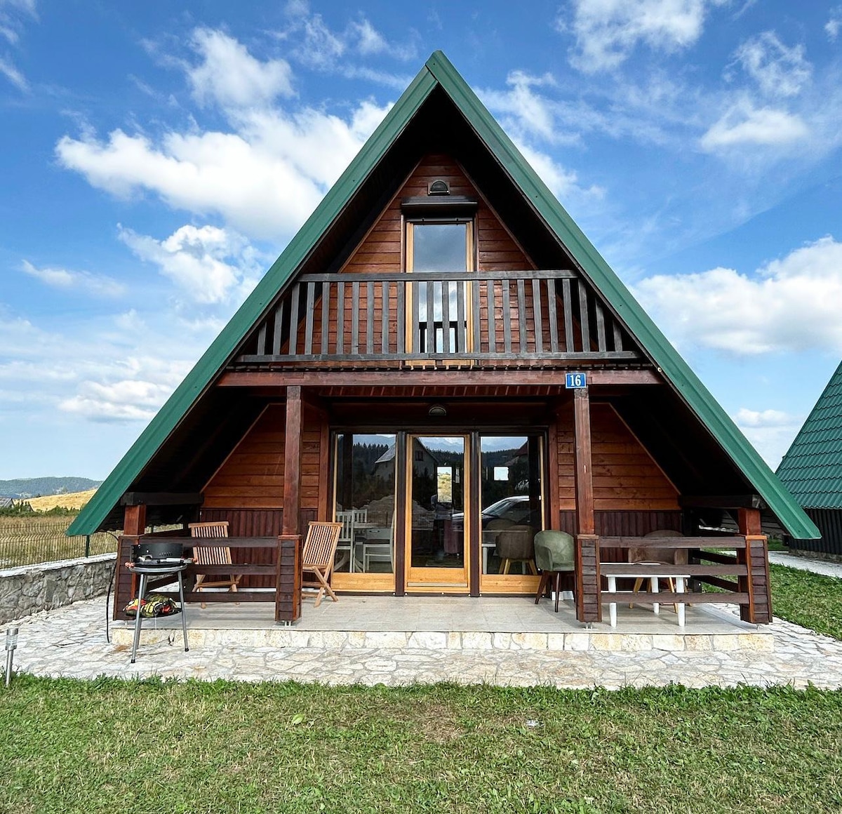 A charming cabin with an A-frame design features a deep green roof and wooden exterior. A spacious porch is furnished with tables and chairs, providing a welcoming entrance. Large glass doors invite natural light, while the surrounding grassy area and distant hills create a serene outdoor setting.