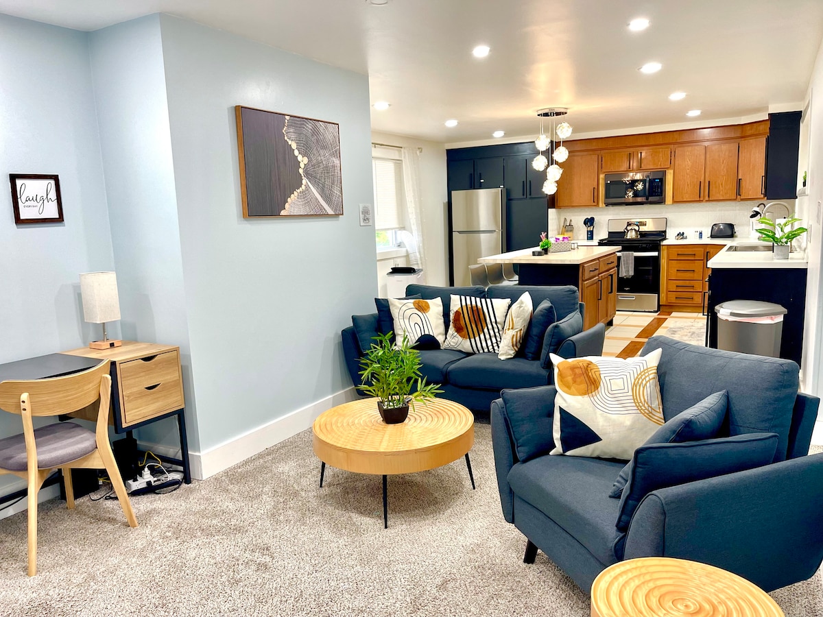 A modern living space features two dark blue sofas adorned with patterned cushions, positioned around a circular wooden coffee table. A sleek desk and chair are visible to one side, while the bright kitchen with wood cabinets and stainless appliances is seen in the background.