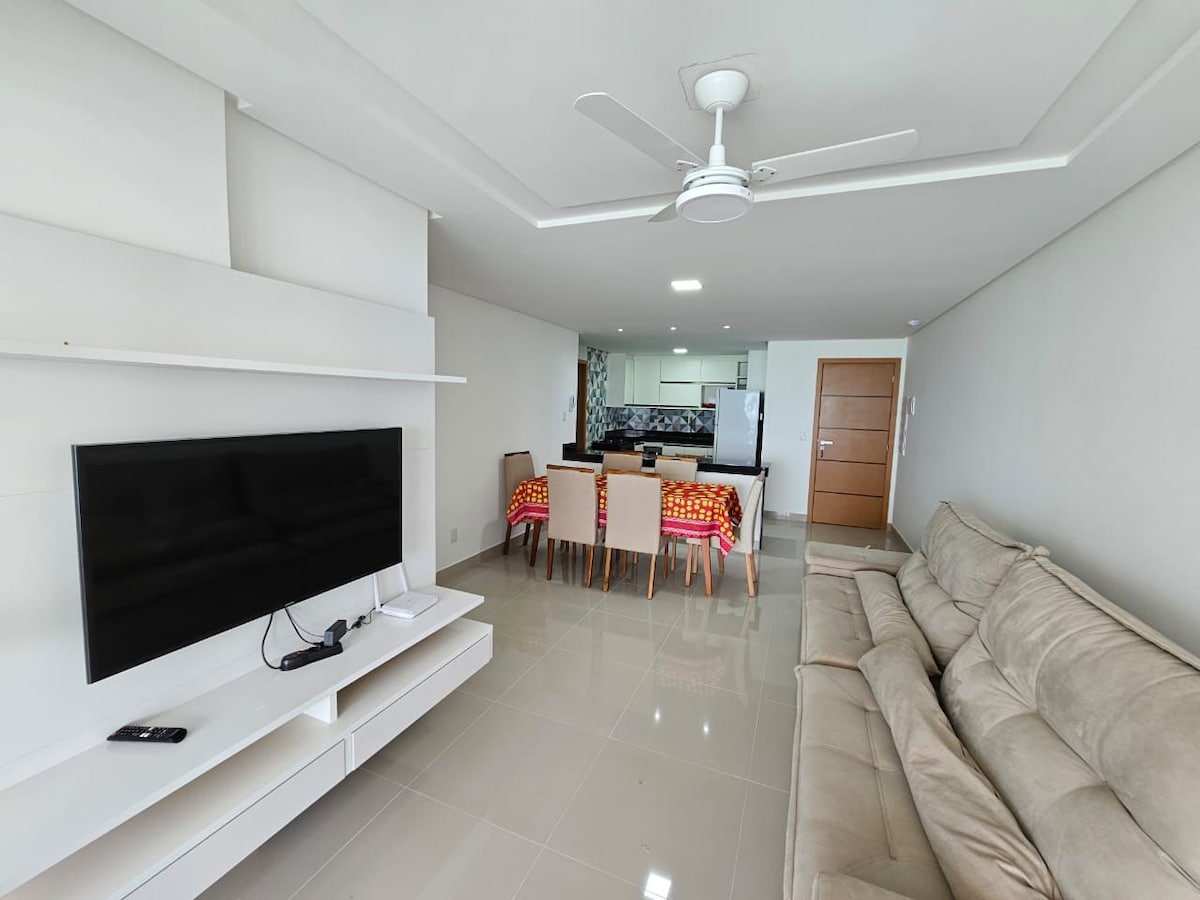 A bright living space features a soft sectional sofa and a mounted television on a white entertainment unit. The dining area, equipped with a large table and colorful chairs, is visible in the background, alongside a well-equipped kitchen. Ceiling fans circulate air throughout the room.