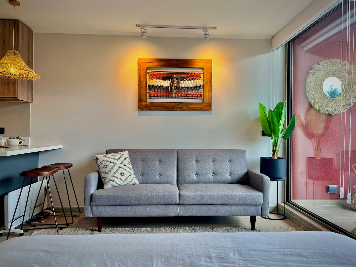 A modern living area features a gray sofa accompanied by a decorative pillow. A framed artwork is displayed on the wall, and a tall green plant adds a touch of nature. Large windows allow for natural light to illuminate the space.