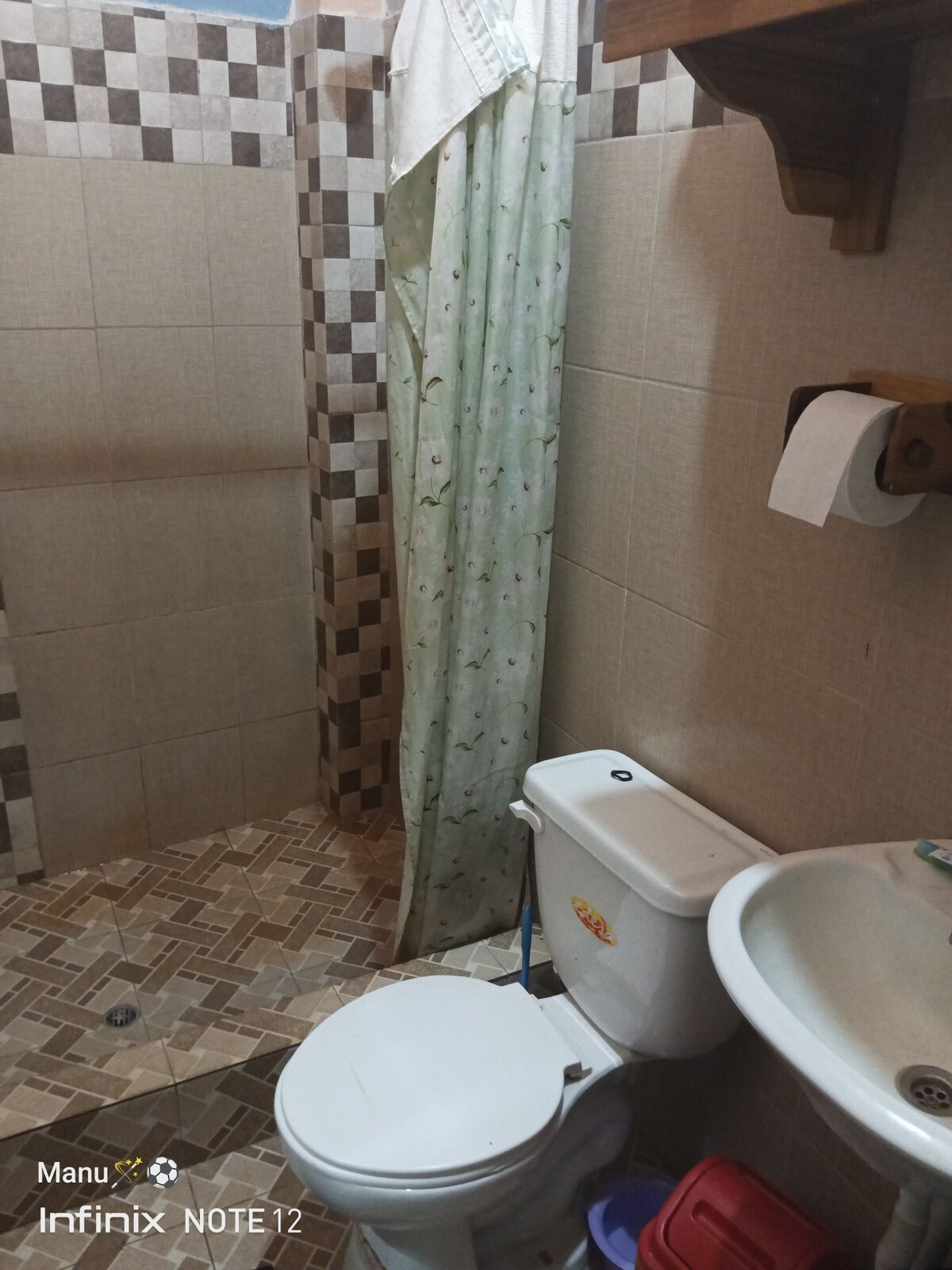 A compact bathroom is equipped with a toilet and a small sink. A shower area features a patterned shower curtain, and tile work covers the floor and walls. Basic toiletries and towel holders are visible.