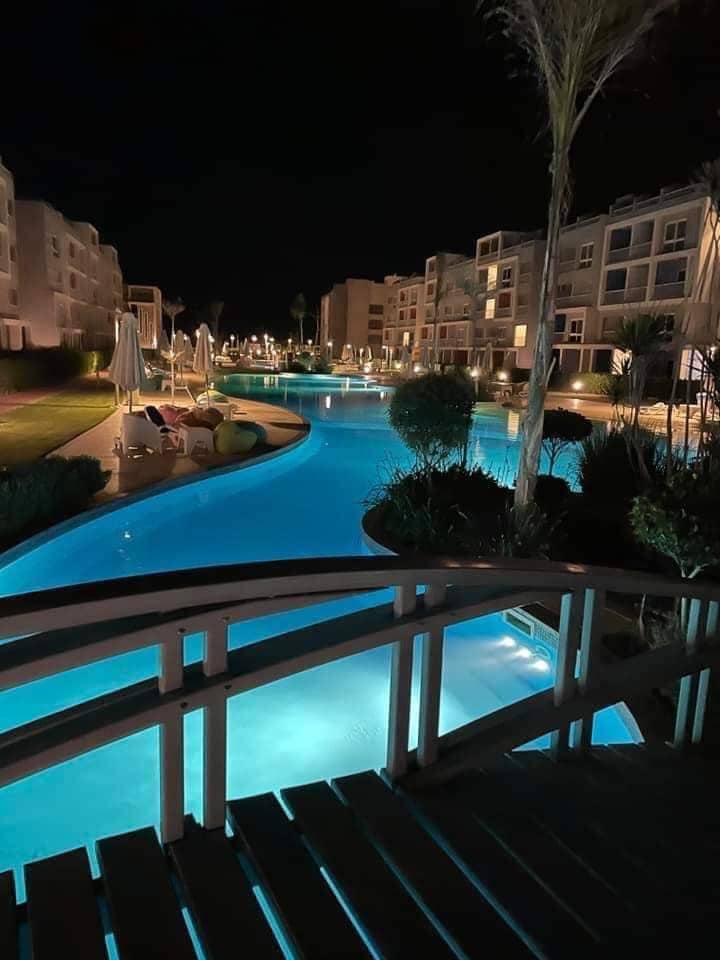 A serene nighttime view of a winding pool, reflecting soft turquoise lights. Lush greenery and palm trees are featured along the edges, complemented by white umbrellas and seating areas. Nearby buildings are softly illuminated, enhancing the peaceful ambiance.