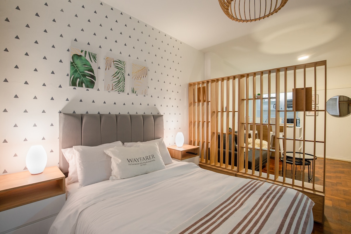 A modern sleeping area features a spacious bed with a light-colored duvet and decorative pillows. Soft lighting is provided by two round lamps on wooden nightstands. A wooden partition adds a level of privacy, while artwork on the wall adds a touch of style.