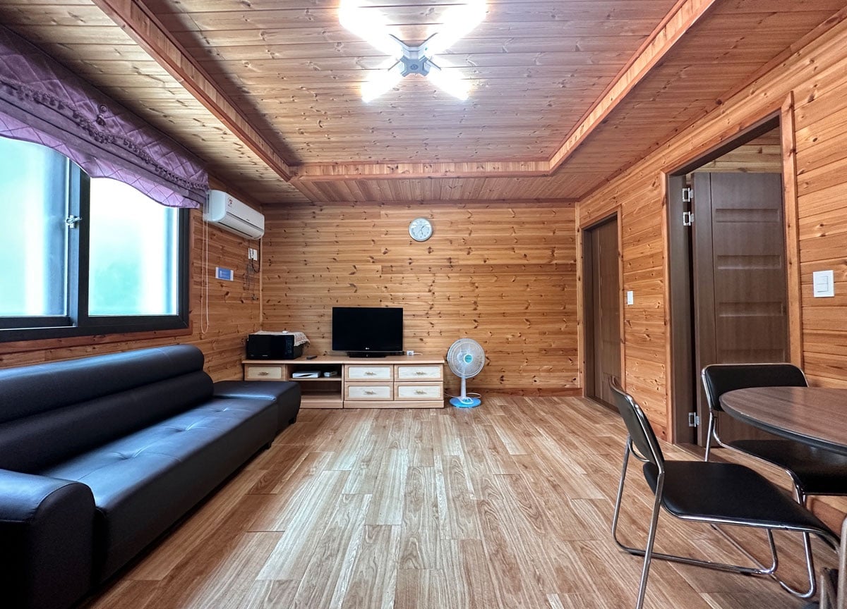 A wooden interior is featured, highlighting smooth paneling and a ceiling fan. A black sofa is positioned alongside a flat-screen TV and a wooden media console. Light-colored flooring enhances the space, which includes a round dining table and chairs, creating a functional living area.