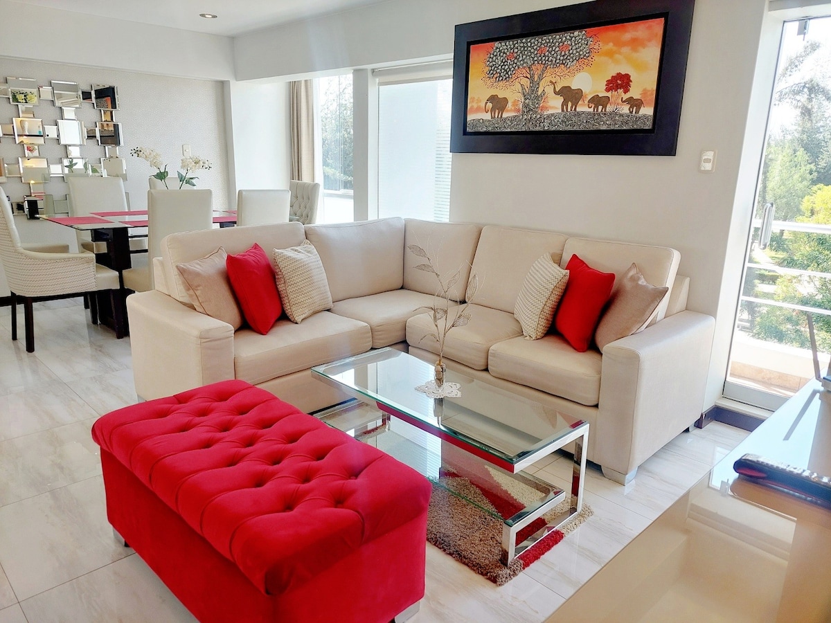 A spacious living area features a light-colored sectional sofa adorned with red accent pillows. A glass coffee table sits on a rug, complemented by a vibrant red ottoman. Large windows provide ample natural light, enhancing the contemporary design of the space.