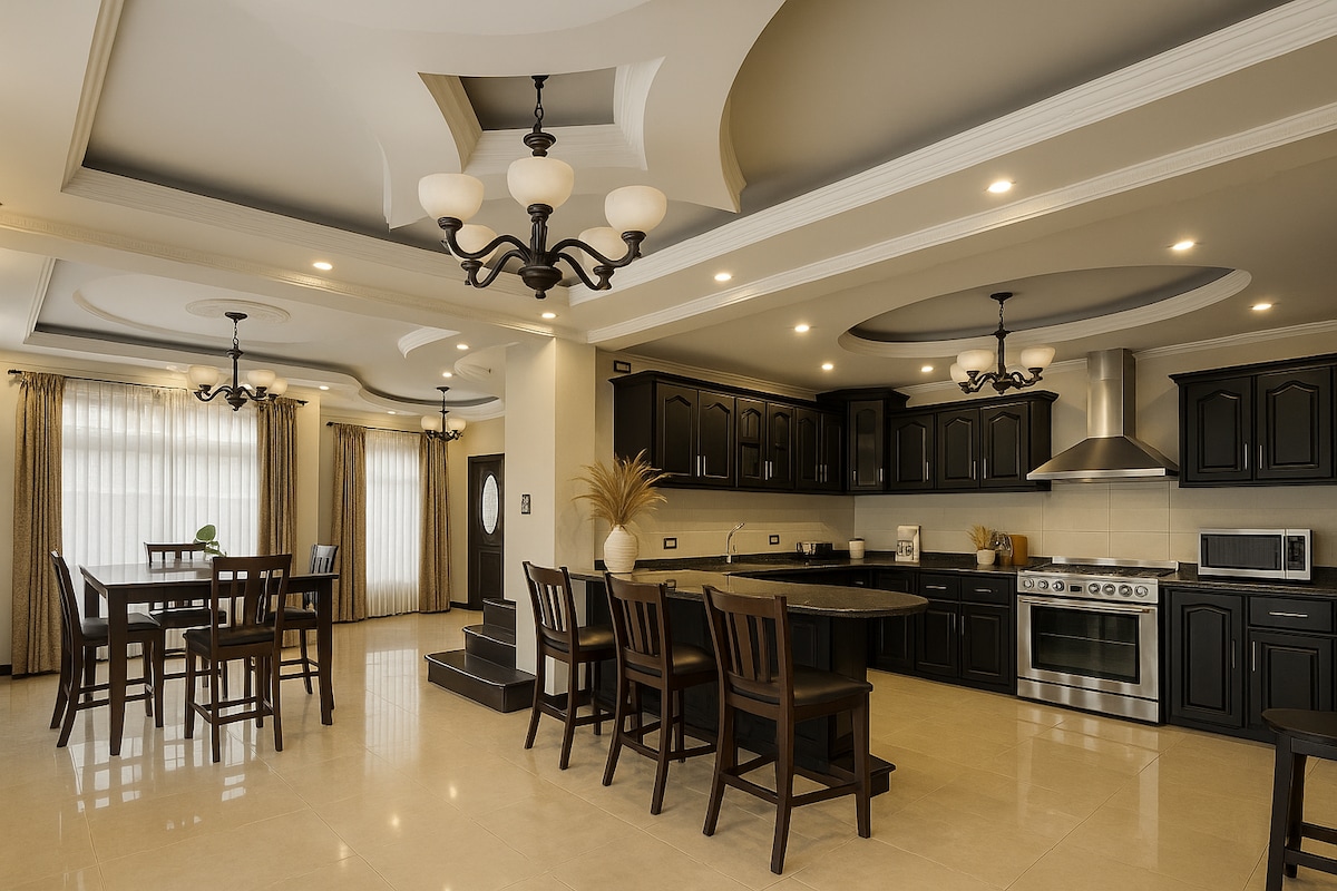 The spacious dining and kitchen area is equipped with dark wood cabinetry and modern appliances. A central island counter provides seating, complemented by a dining table featuring four chairs. Large windows allow natural light to fill the room, accented by decorative ceilings and elegant light fixtures.
