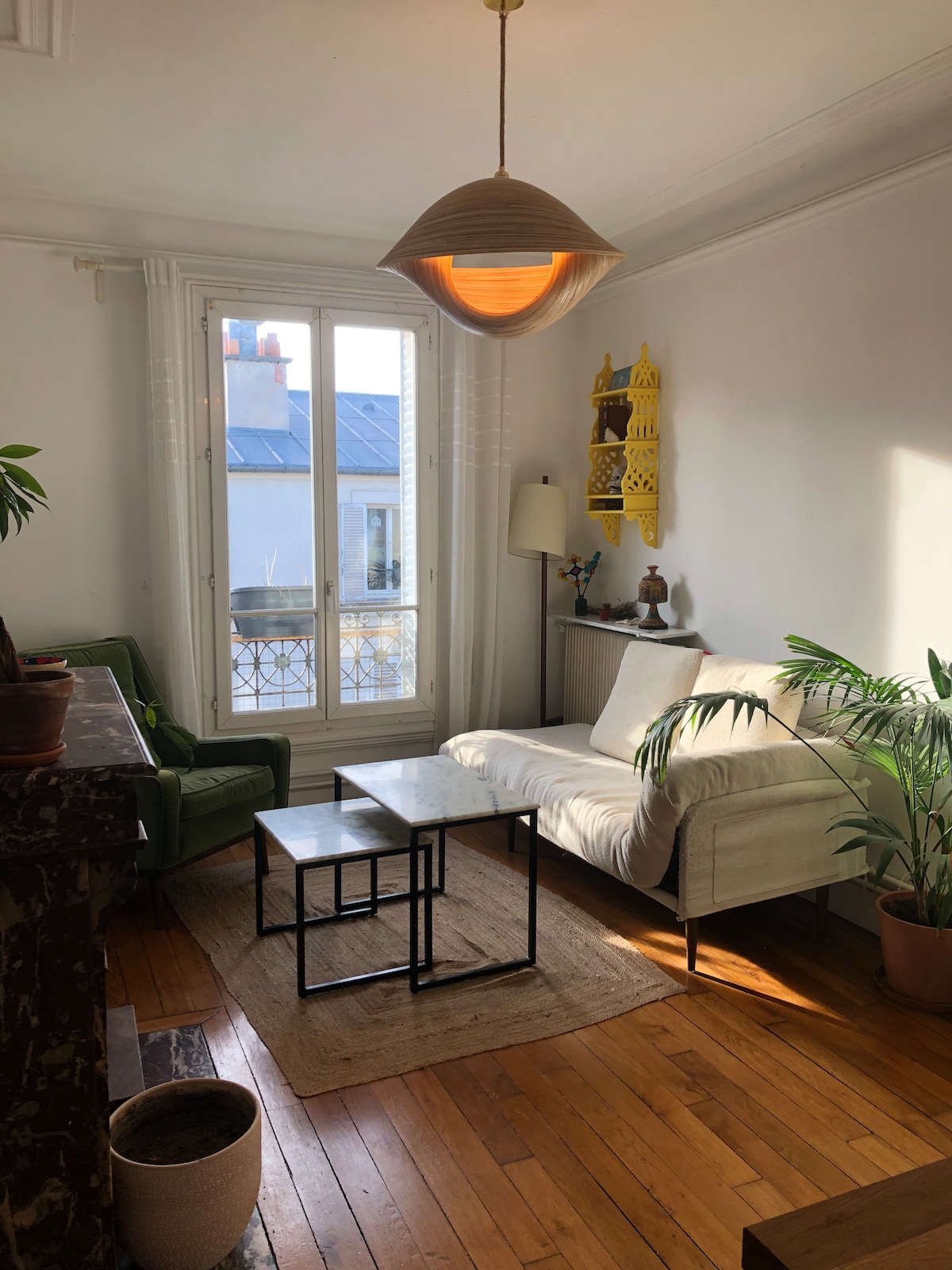 A cozy living area features a light-colored sofa and two green chairs, accented by a warm wooden floor. Two black tables are positioned on a neutral rug. Sunlight streams through large windows with white shutters, illuminating the room and highlighting an artistic wall decor.