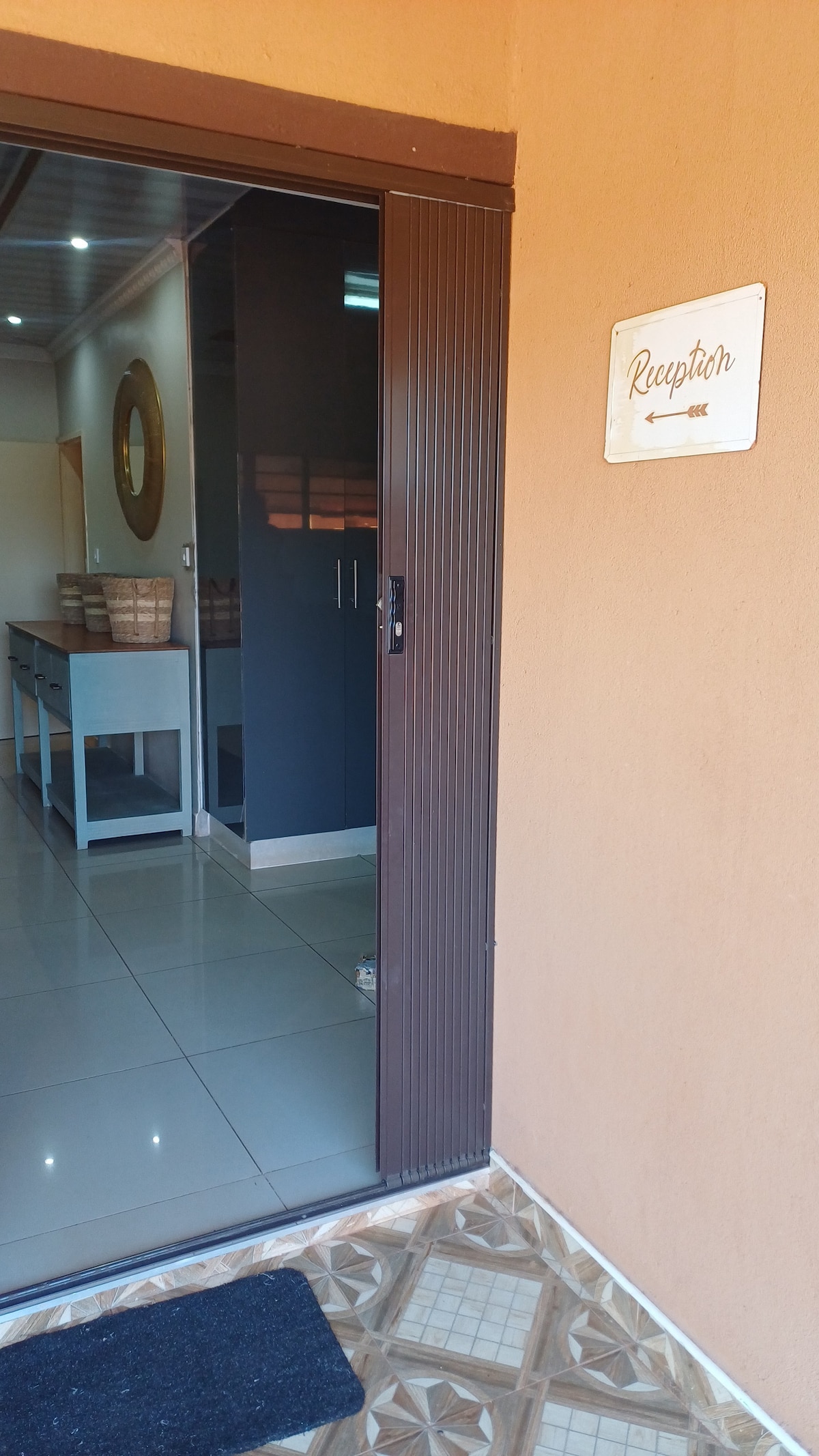 A welcoming entrance is marked by a sign indicating 'Reception' with an arrow. The polished tile floor leads to a reception area, featuring a round mirror and a wooden table with a decorative basket. Soft lighting enhances the space's clarity.