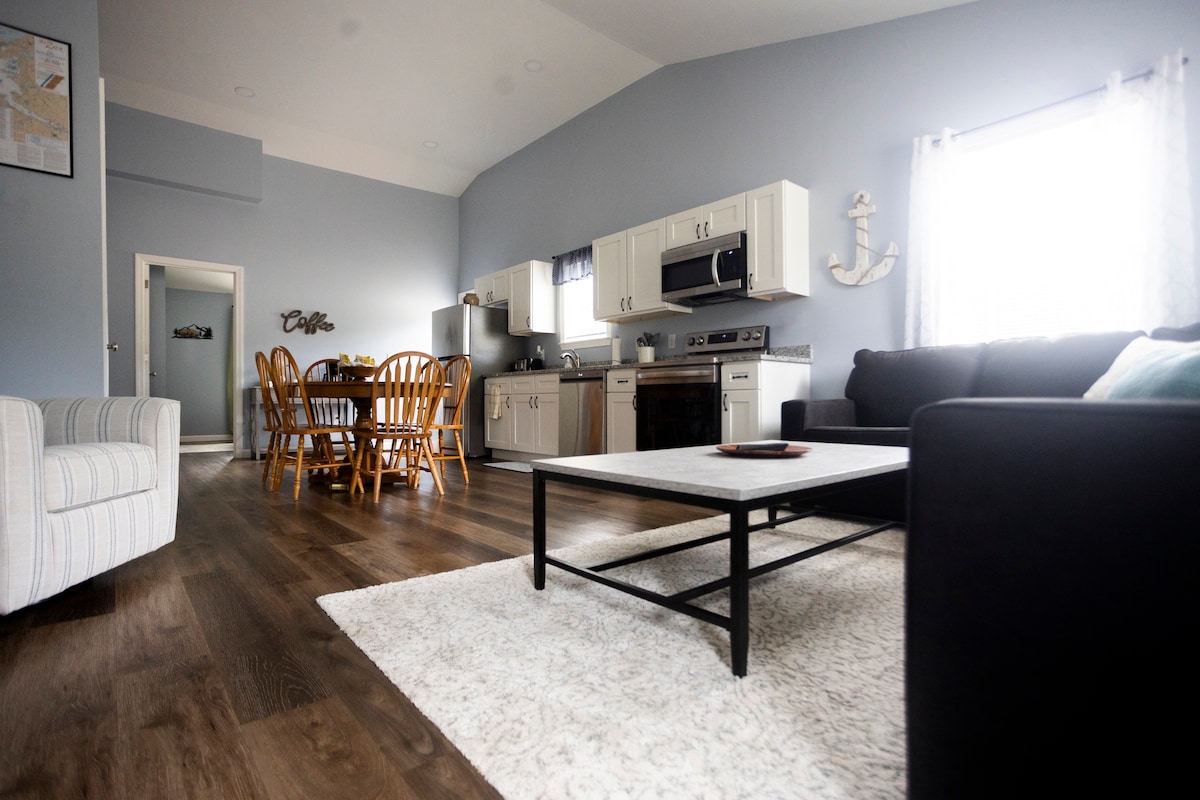An open living space features a modern kitchen with stainless steel appliances and white cabinetry. A dining table with six chairs is visible, and a cozy seating area is arranged around a coffee table on a soft rug. Natural light enters through a window adorned with light curtains.