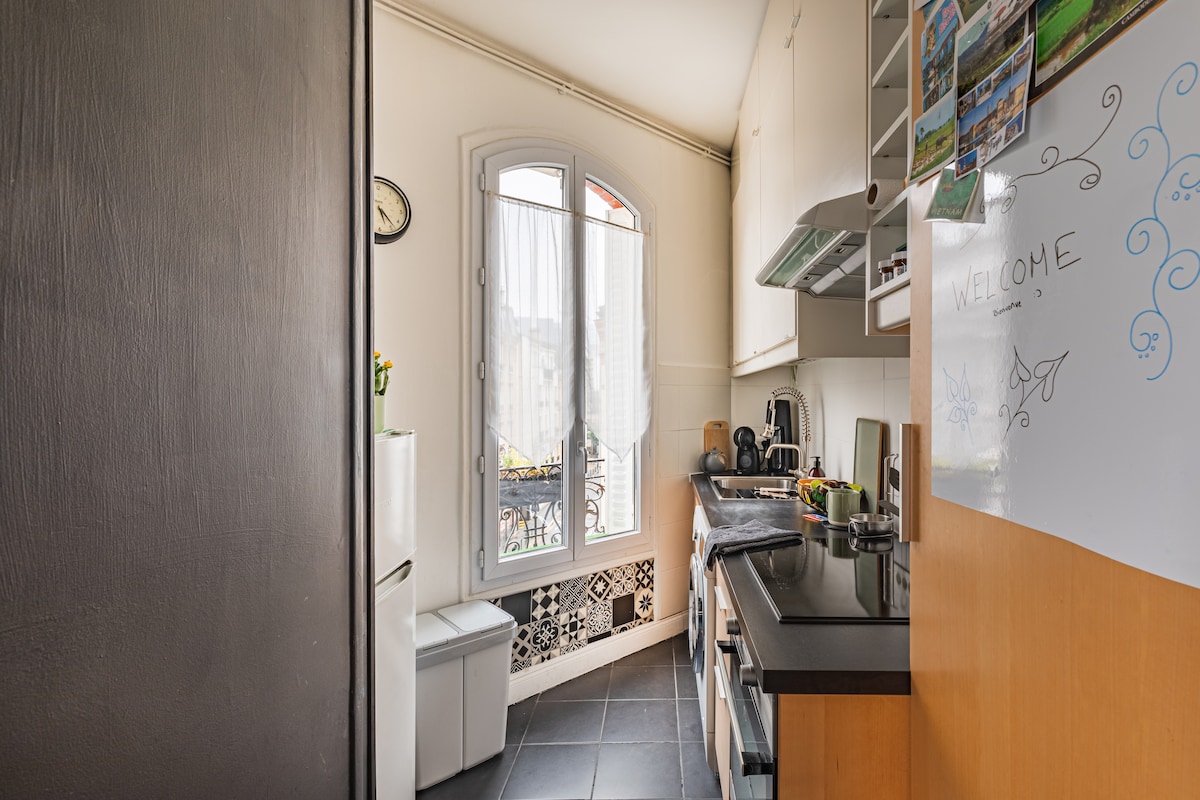 A compact kitchen features modern appliances, including a stove, oven, and refrigerator. Natural light enters through a window with light curtains, enhancing the bright atmosphere. Sleek cabinetry provides storage solutions, while a chalkboard adds a personal touch. The floor is tiled in dark tones.
