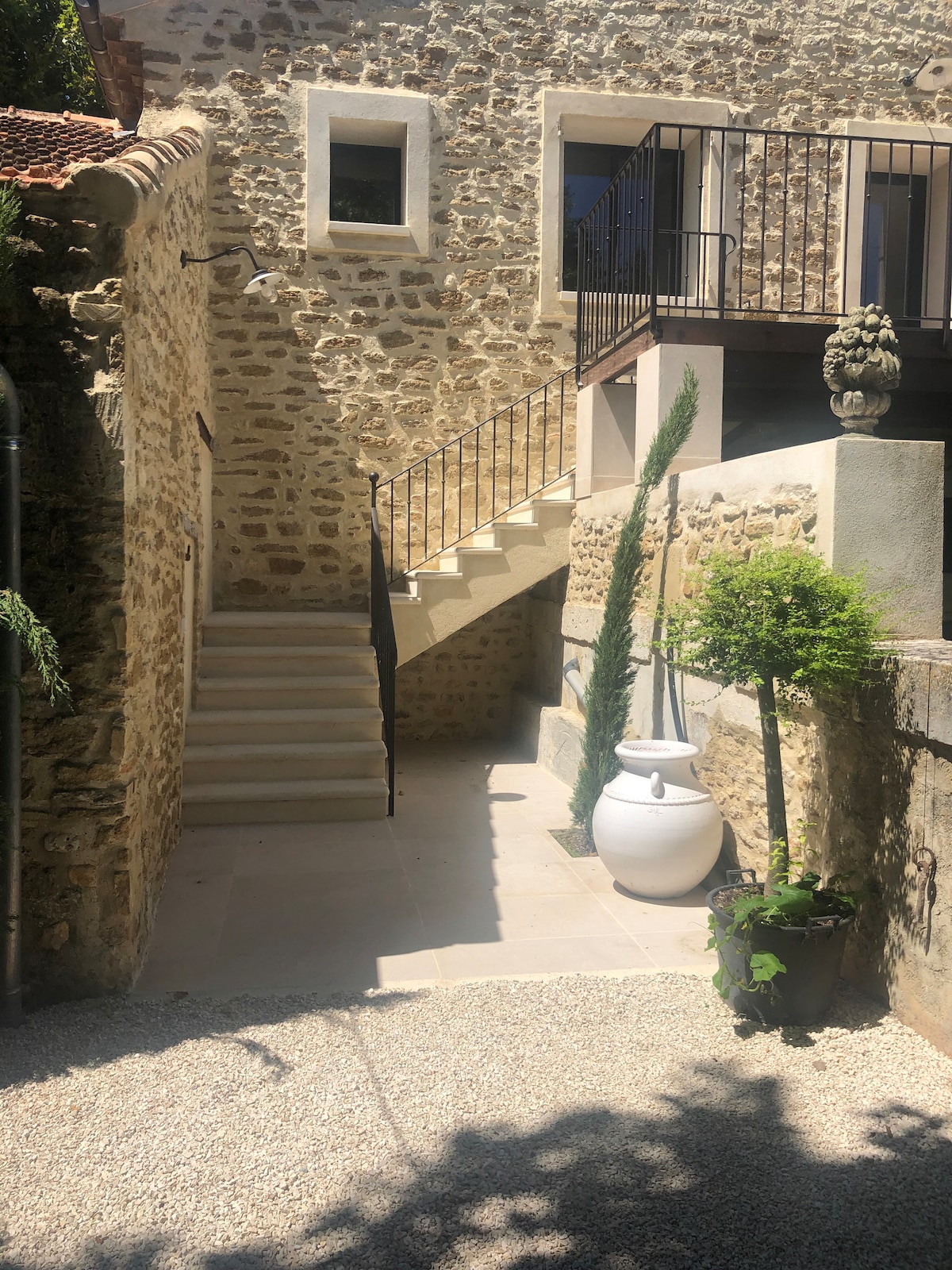An outdoor area is depicted, featuring a stone wall structure with a staircase leading to an upper level. Decorative plants flank the base of the stairs, and a large white pot is positioned on a smooth stone surface, adding a touch of greenery to the entrance.