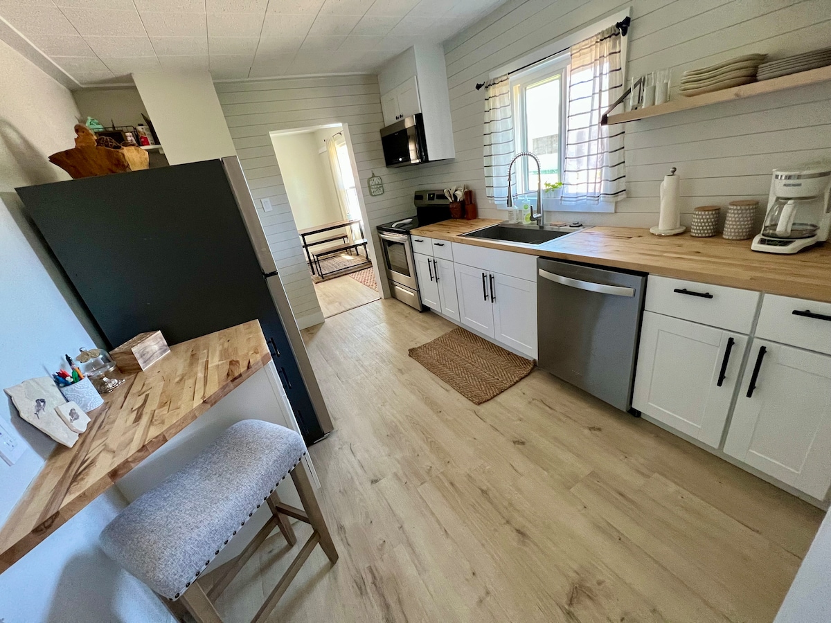 The kitchen features modern appliances and wooden countertops. A stainless steel sink is positioned under a window, while white cabinets offer ample storage. A small dining area is included with a cushioned stool, and light-colored flooring enhances the spacious feel of the room.