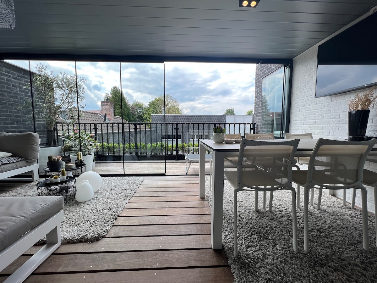 A modern outdoor space features a wooden deck with a white dining table surrounded by four chairs. A comfortable seating area is accentuated by soft cushions. Floor-to-ceiling glass walls offer views of the greenery beyond, allowing natural light to fill the area.