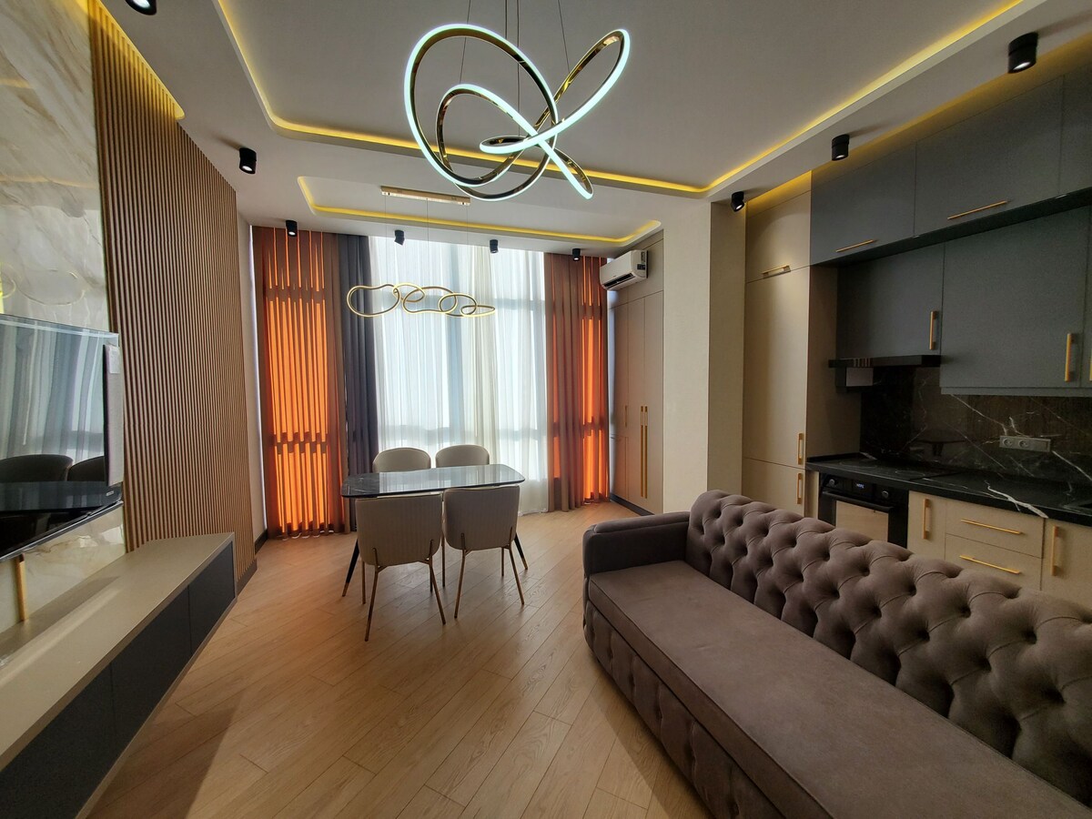 A modern living space is presented with sleek furnishings. A sectional sofa in a neutral tone complements a stylish dining table surrounded by three chairs. Bright orange and gentle white curtains frame a large window, while a contemporary light fixture adds interest to the ceiling.