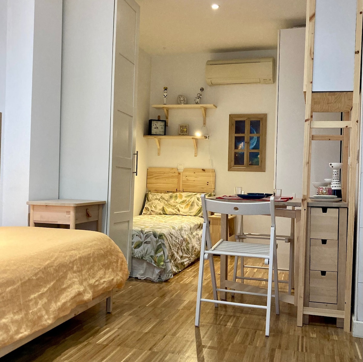 A spacious room features a queen-sized bed covered in soft beige bedding. A smaller bed with floral patterns is visible in the background. Wooden furniture includes a table and chairs, with shelves and artwork adorning the walls, contributing to a cozy, functional layout.