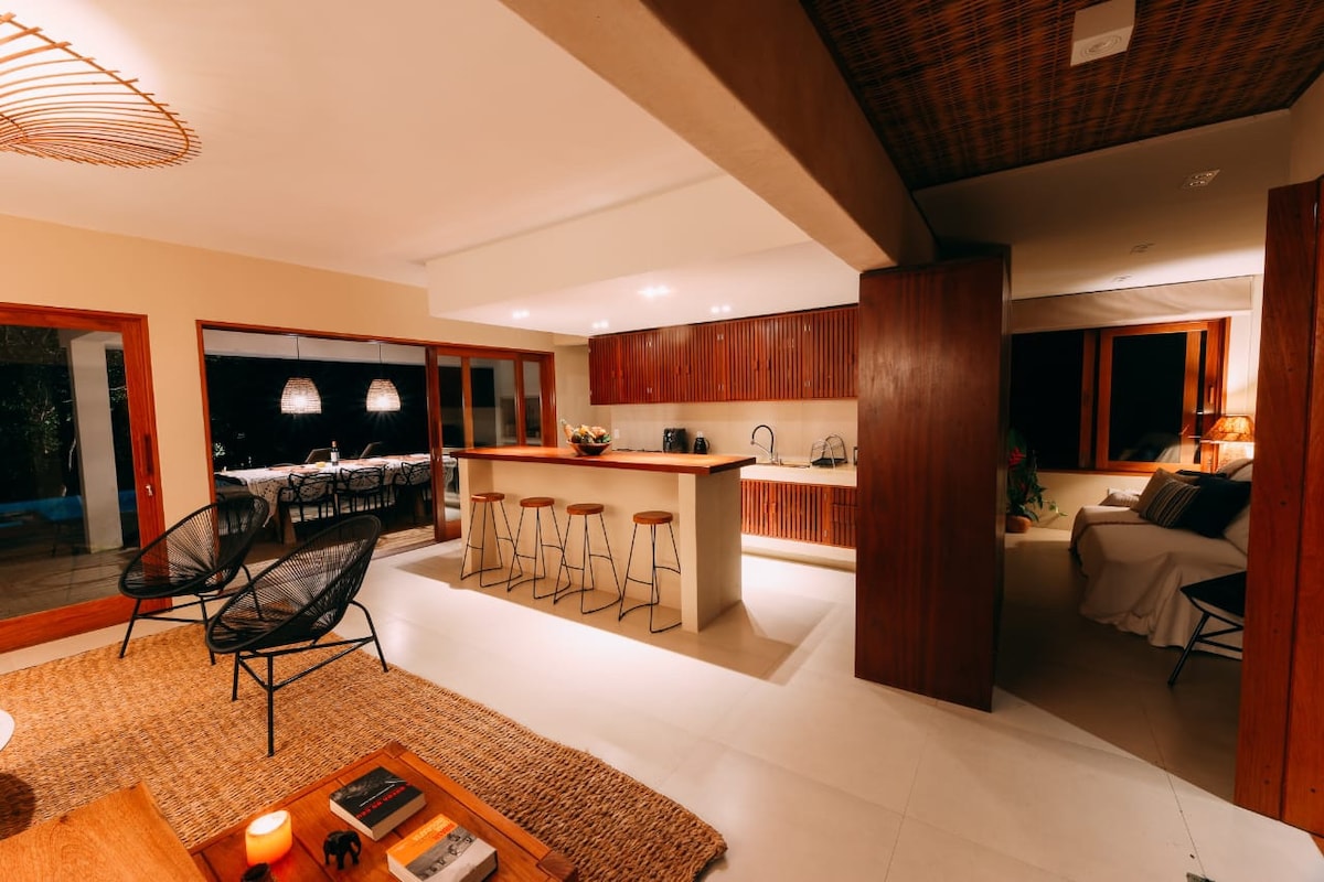 The open-concept living space features a modern kitchen with wooden cabinetry and a large island. Two lounge chairs are positioned nearby on a natural fiber rug, while large windows frame views of the outdoor area. Warm lighting creates a cozy ambiance throughout.